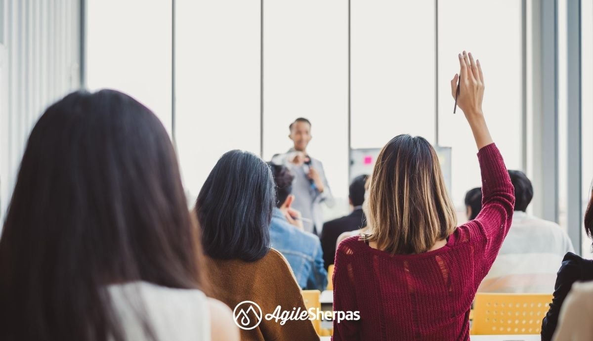 A person standing next to a Kanban board taking questions from an audience in front of him.